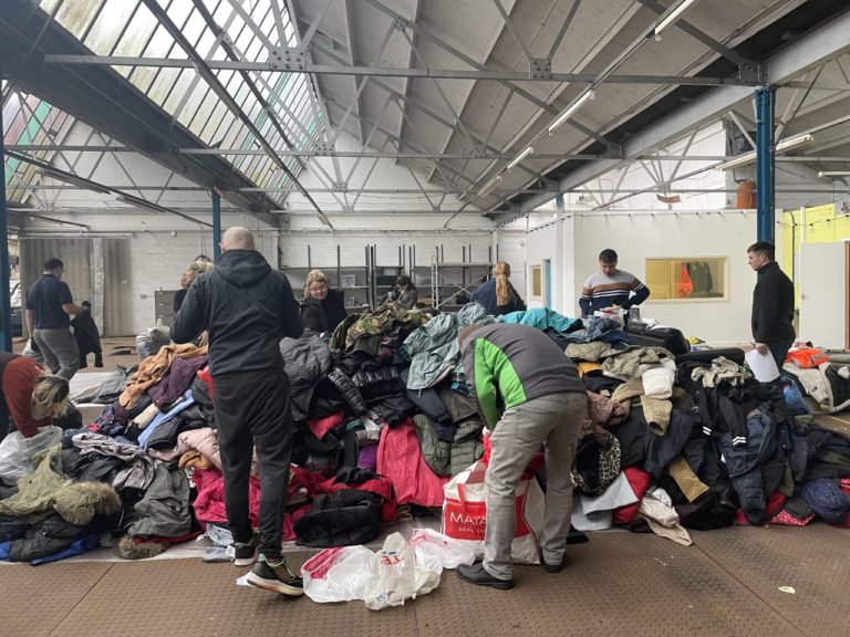 An image of a pile of coats on the floor of a warehouse with individuals putting more coats on the pile