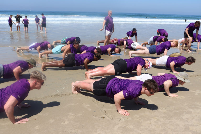 An image of rugby players doing push ups, in purple shirts, on a beach