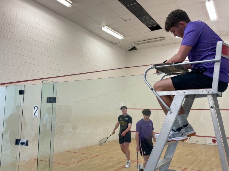 A man in a high chair overlooking a squash court, keeping track of the score. Two man, one in a purple top and one in a black top, in the Squash Court playing against each other.