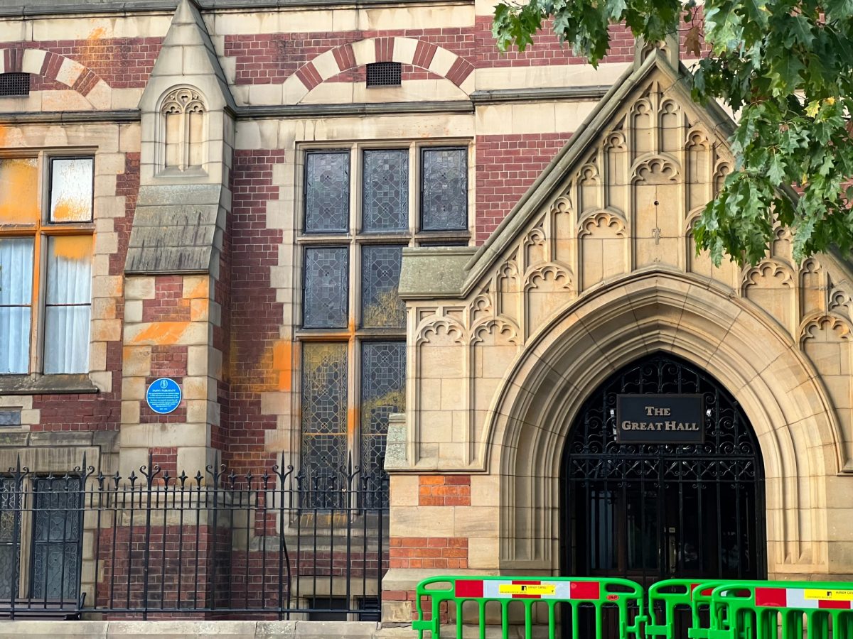 Image shows the entrance to Leeds University building, The Great Hall. Orange paint is splashed across the windows to the left of the entrance and green barriers have been placed to block the buildings entrance.