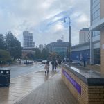 The image shows a Leeds Becket University sign with standing water on top. The city of Leeds is in the background with high rise buildings and people walking in the street.