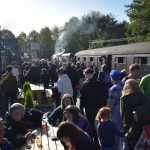 Crowds on people getting off an old steam train at Oxenhope station. There are people sat at a picnic bench on the left. The sun is shining and the sky is blue