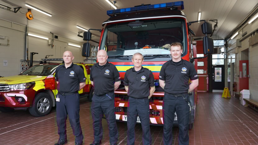 four Fire fighters stood in front a fire engine at a fire station