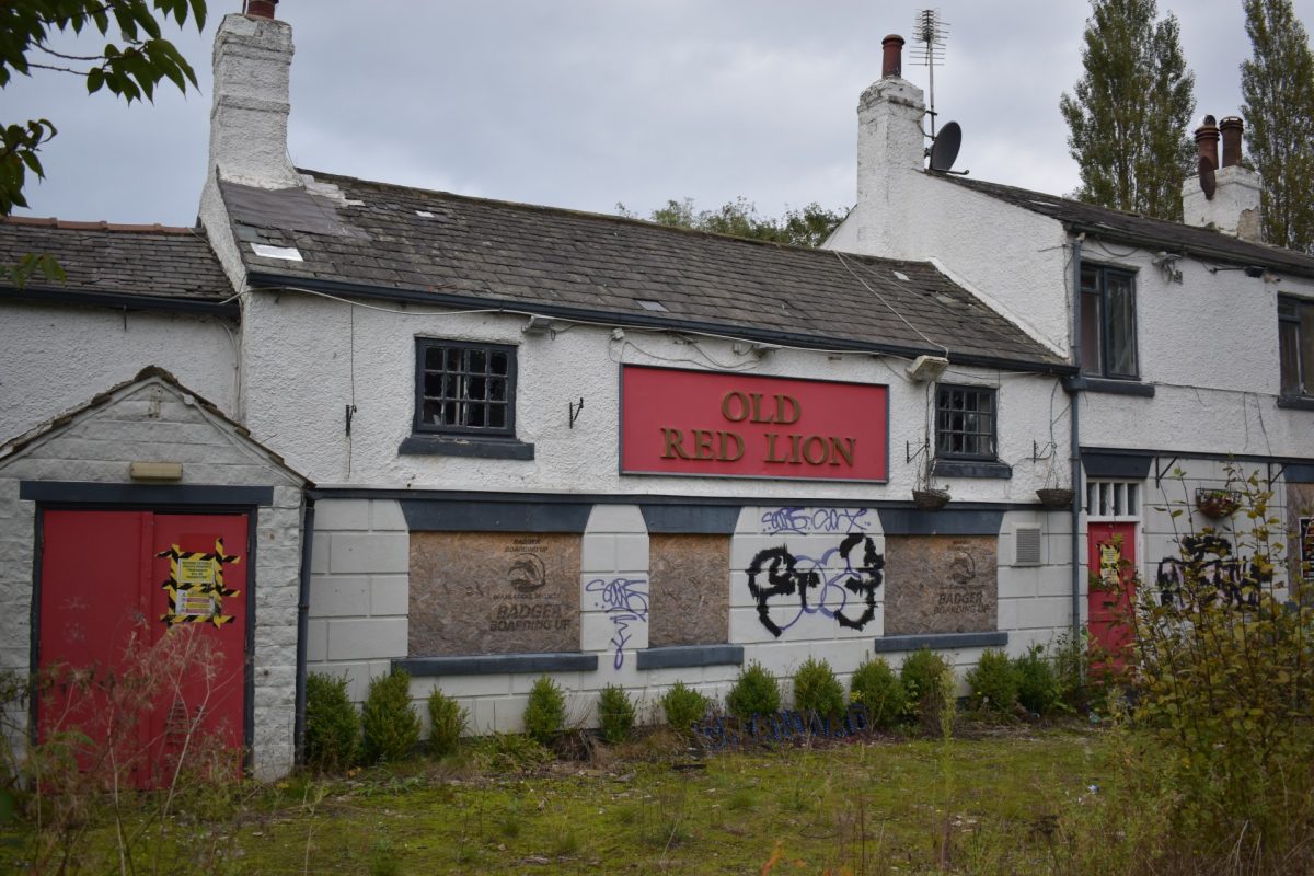 An image of the Old Red Lion pub located on York Road, Whinmoor. The building is white with red signage. The bottom windows are boarded up and the top windows have been smashed. The grass at the front of the building is overgrown.