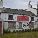 An image of the Old Red Lion pub located on York Road, Whinmoor. The building is white with red signage. The bottom windows are boarded up and the top windows have been smashed. The grass at the front of the building is overgrown.