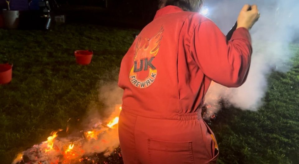 This image depicts a UK Firewalk worker wearing a red jumpsuit that says 'UK Firewalk'. She's shovelling the coals ready for people to walk across.