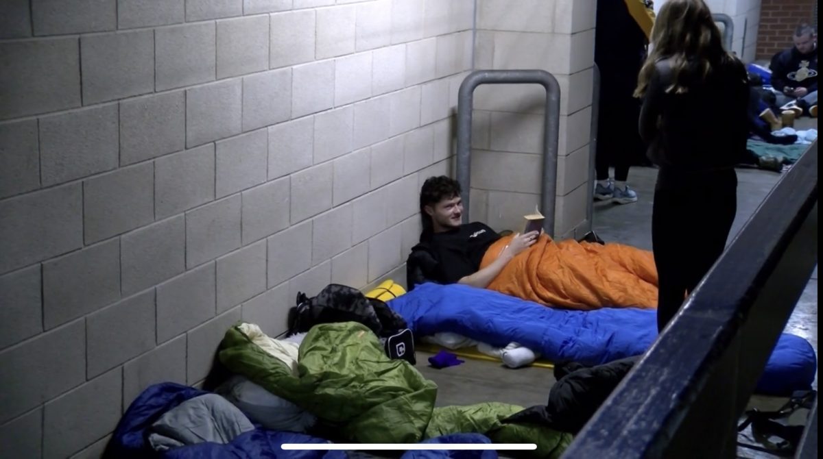 A man is laying on the floor of the Headingley Stadium stands with a book in his hands in an orange sleeping bag as he prepares to sleep