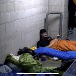 A man is laying on the floor of the Headingley Stadium stands with a book in his hands in an orange sleeping bag as he prepares to sleep