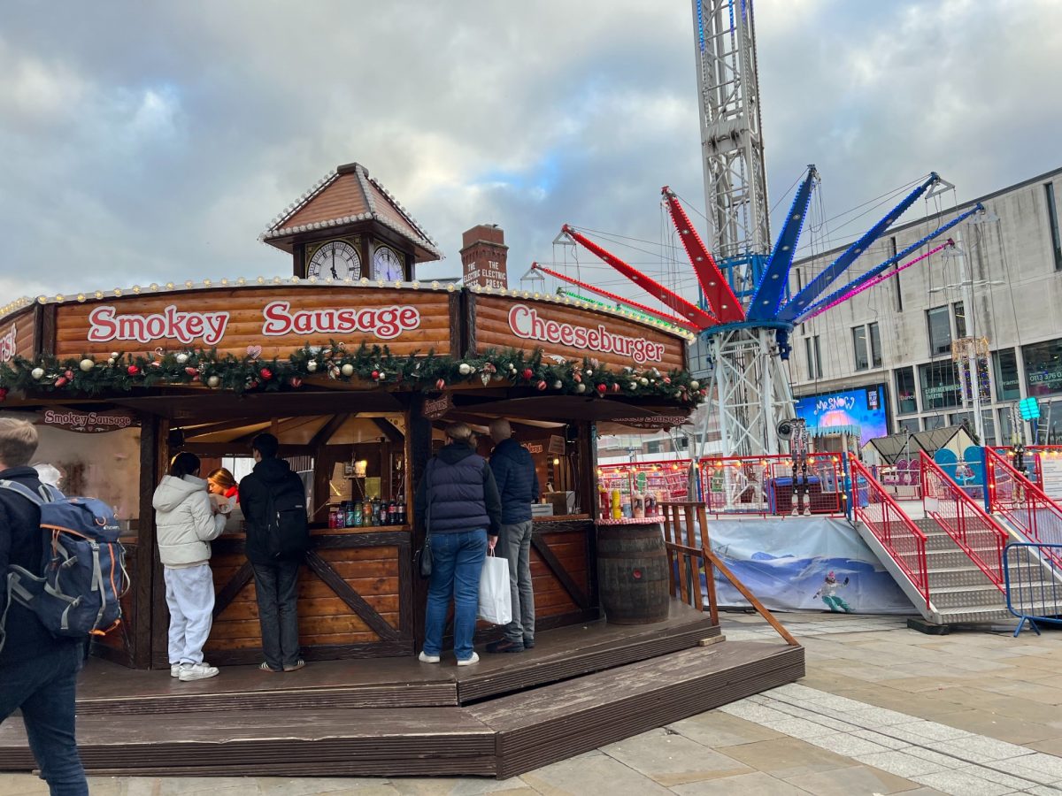 This image depicts a round,, wooden German sausage stand at Leeds Christmas markets where people can be seen queueing. There is a fairground ride behind the stand.
