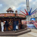 This image depicts a round,, wooden German sausage stand at Leeds Christmas markets where people can be seen queueing. There is a fairground ride behind the stand.