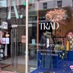 This image shows the glass frontage of the Trad Collective store on the high street in Leeds. There is a woman with pink hair walking in front of the window