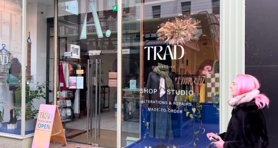 This image shows the glass frontage of the Trad Collective store on the high street in Leeds. There is a woman with pink hair walking in front of the window