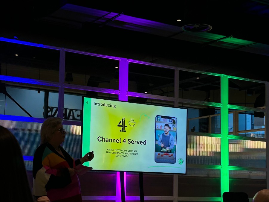 A person stands presenting in front of a screen displaying Channel 4 Served, with colourful lights in the background and an image of a man cooking on the screen.
