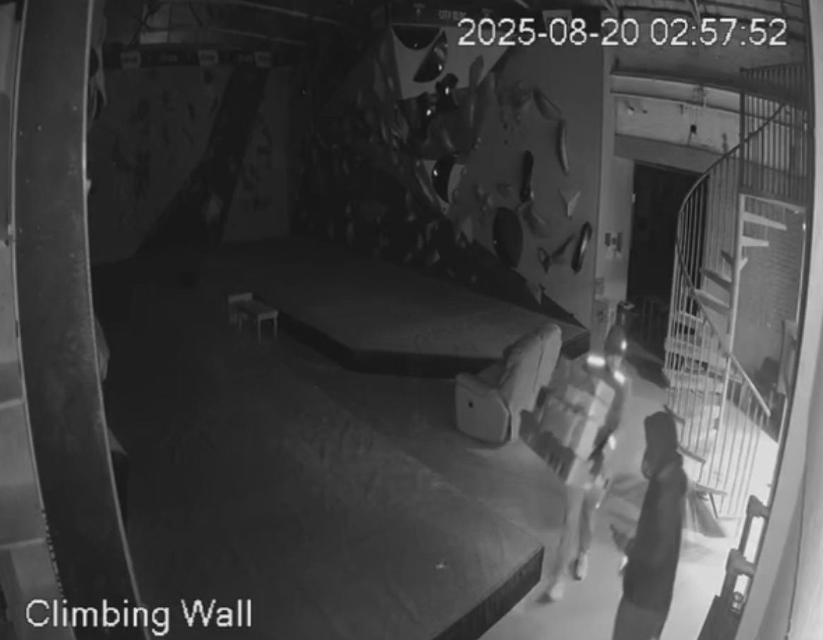 Security camera footage from a climbing gym dated 2025-08-20 at 02:57:52 AM. The image shows two individuals in the lower right corner; one is carrying a large box and wearing a reflective vest, while the other is in a hoodie. The climbing wall is dimly lit in the background, and a spiral staircase is visible on the right side of the image. The words “Climbing Wall” are labeled at the bottom left of the frame.