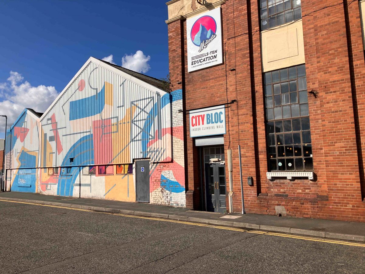 Street view of a colourful building with geometric mural art on its corrugated metal exterior. The mural features abstract shapes in red, blue, yellow, and peach tones. Attached to the building is a red-brick structure with tall industrial-style windows. Two signs are visible: one reads “CITY BLOC Indoor Climbing Wall” above a door, and the other above it reads “LS-TEN Education” with a mountain logo. The sky is clear and blue with scattered clouds.