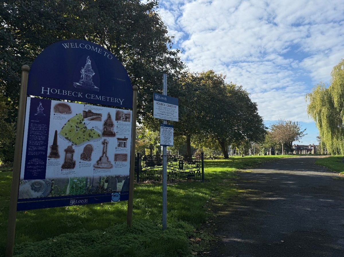 A bright, slightly overcast day at Holbeck Cemetery in Leeds, England. The foreground features a large, dark blue welcome sign with white text reading "Welcome to Holbeck Cemetery," accompanied by a detailed map and illustrations of notable monuments within the grounds. Beneath the map, the sign is labeled with the Leeds City Council logo. Nearby, a smaller white sign also displays the cemetery's name and some rules or information for visitors.

To the right of the signs, a paved path winds into the cemetery, flanked by green grass and mature trees, including a large willow tree. The cemetery itself stretches into the background with visible headstones and monuments scattered among the trees. The sky is mostly blue with patches of fluffy white clouds, creating a peaceful and serene atmosphere.