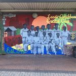 A mural on a brick wall titled “Leeds Caribbean Cricket Club,” depicting a historic black-and-white photo of a cricket team composed of Black Caribbean men. The background is vibrant with a sunset, palm trees, and Caribbean huts.