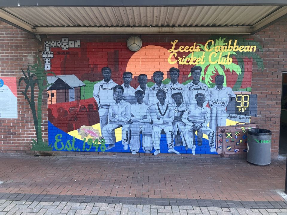 A mural on a brick wall titled “Leeds Caribbean Cricket Club,” depicting a historic black-and-white photo of a cricket team composed of Black Caribbean men. The background is vibrant with a sunset, palm trees, and Caribbean huts.
