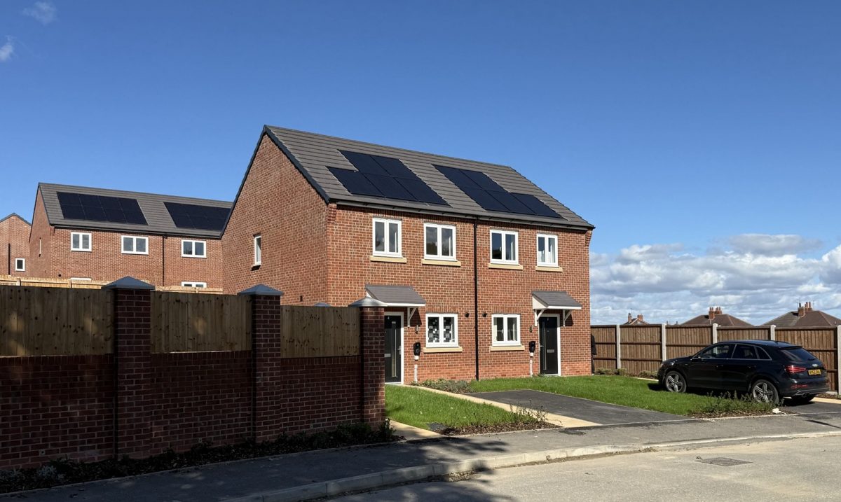 A pair of modern semi-detached brick houses under a clear blue sky, each with solar panels installed on the roof. The homes have white-framed windows and black front doors with small awnings above them. A short path leads to each entrance, and one of the homes has a driveway with a black car parked on it. A mix of wooden and brick fencing encloses the property, and similar houses are visible in the background.