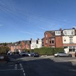 A residential street with brick houses, parked cars, greenery, utility poles, and bins under a clear blue sky.