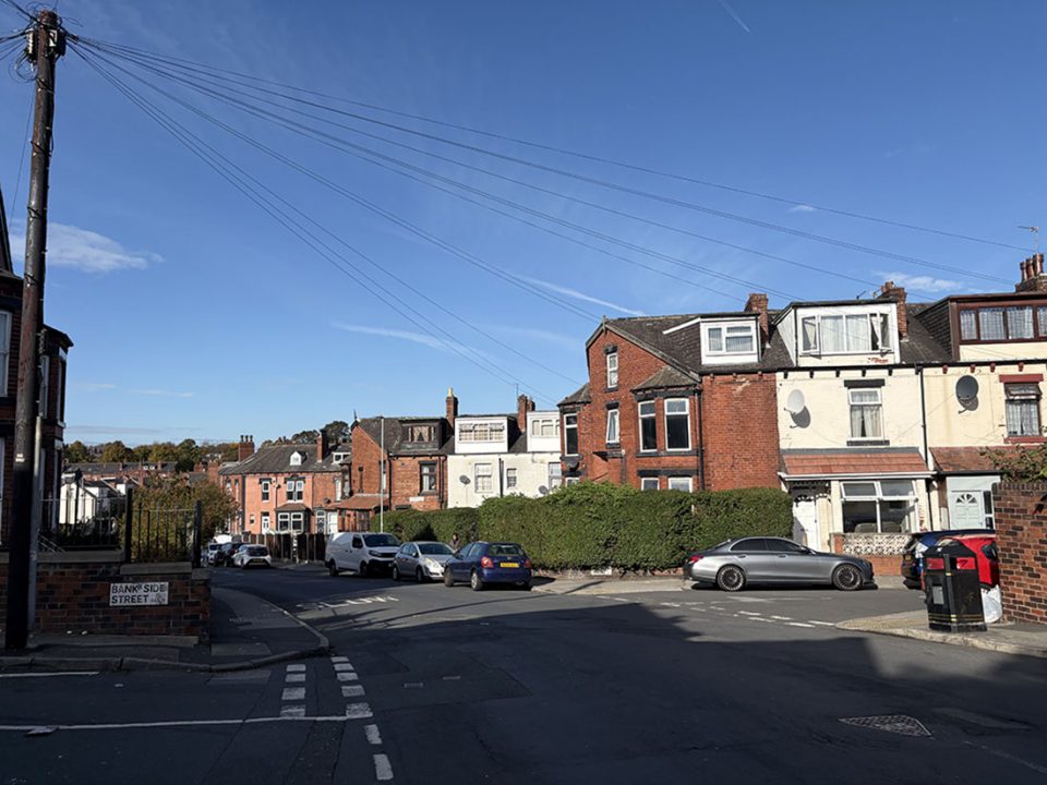 A residential street with brick houses, parked cars, greenery, utility poles, and bins under a clear blue sky.