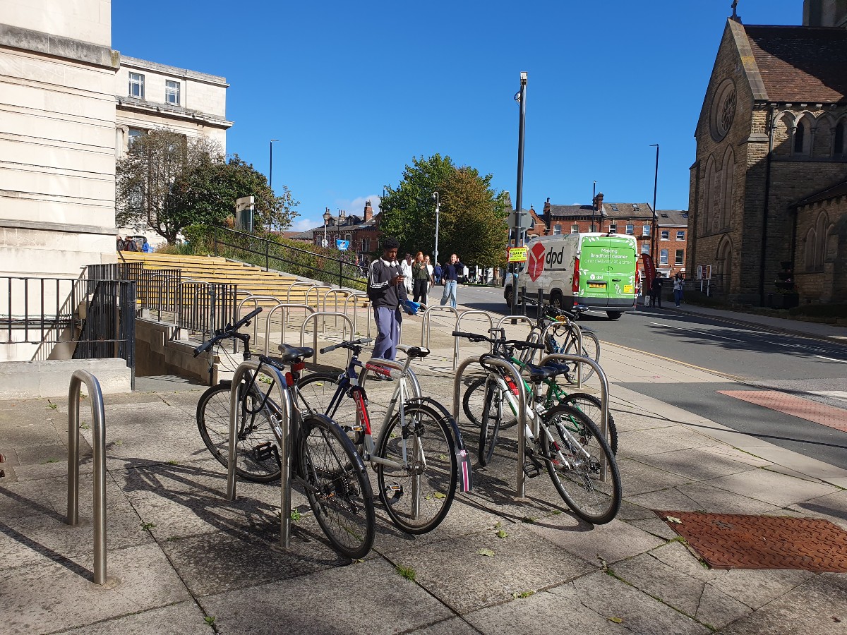 The image shows a bike rack in the University of Leeds campus facing Woodhouse Lane, with pedestrians and a van in the background