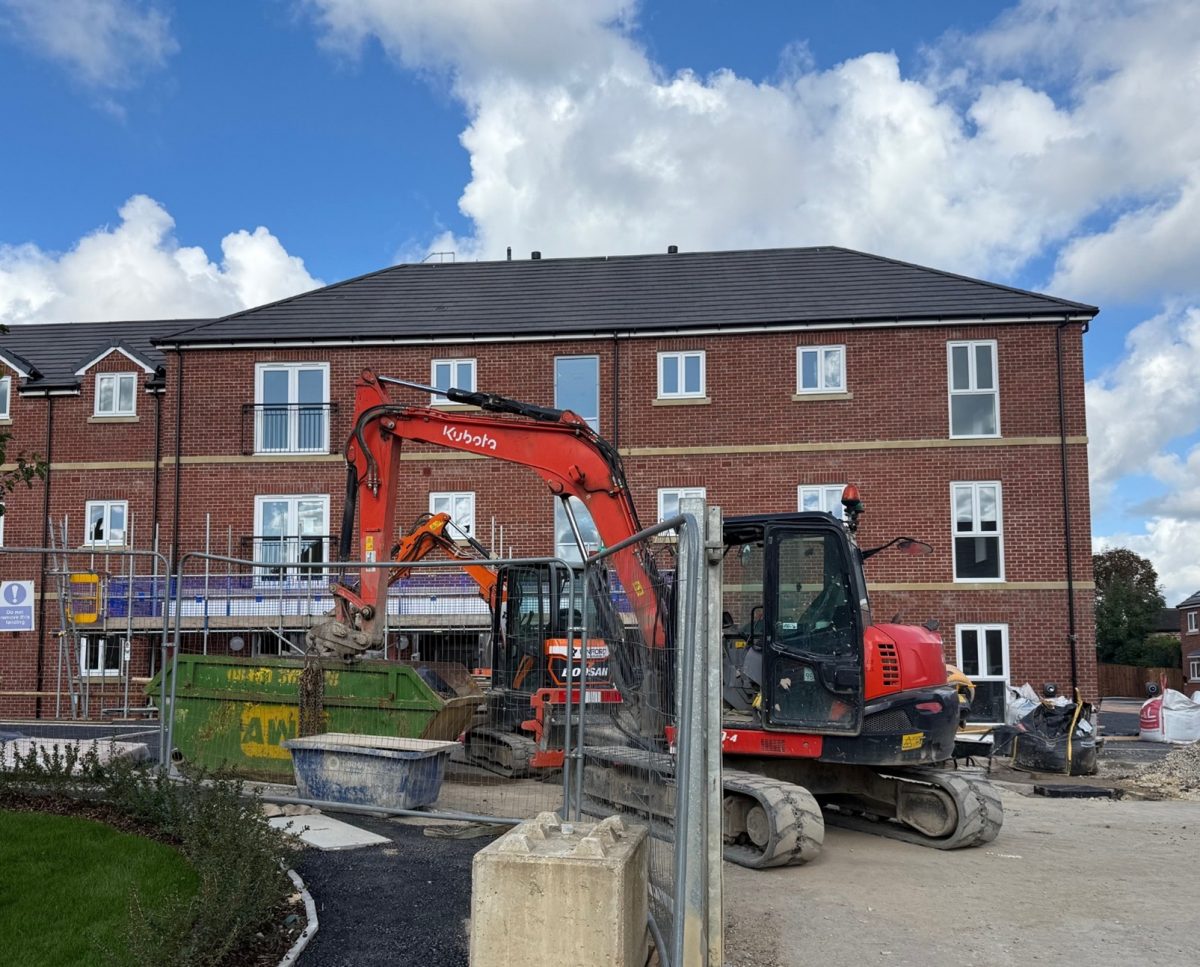A construction site in front of a newly built three-story red-brick apartment building with white-framed windows and glass Juliet balconies. In the foreground, two orange excavators, one branded "Kubota" and the other "Doosan," are actively moving materials into a large green skip. The area is enclosed by metal safety fencing, with construction equipment, materials, and waste bags scattered around. A curved path with fresh landscaping borders the work area, while a partly cloudy sky creates a bright backdrop.