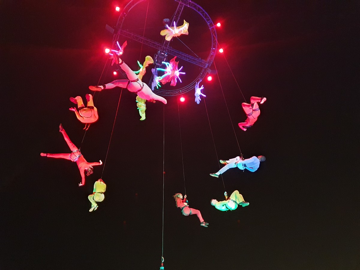 The image shows a number of acrobats performing in the sky attached to a crane