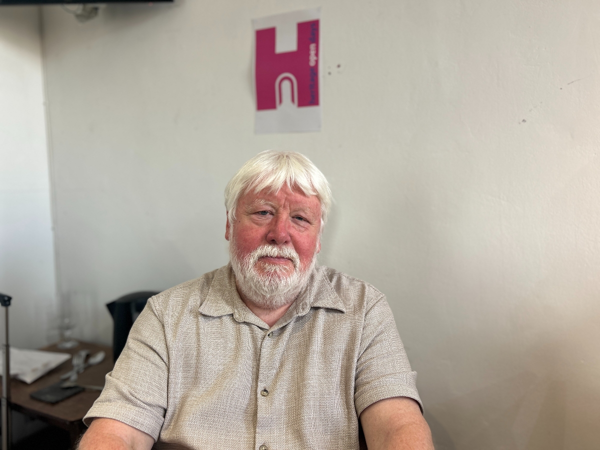 David Iredale - An older man with white hair and a white beard sits indoors against a white wall. He wears a light-colored, short-sleeved, patterned button-up shirt. Behind him is a table cluttered with papers, a black pitcher, and a glass. A poster with a stylized pink and white design featuring the letter "H" hangs on the wall. The setting appears casual, possibly an office or meeting room.