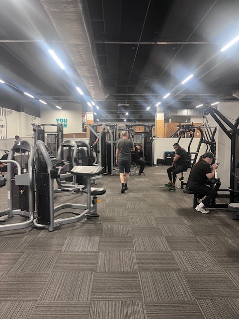 Modern gym interior with various exercise machines and equipment. Several people are present: one walking down the central aisle, two seated on benches using their phones, and others actively working out. The space features bright overhead lighting, gray flooring, and black ceiling panels.