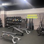 Modern gym interior with various exercise machines and equipment. Several people are present: one walking down the central aisle, two seated on benches using their phones, and others actively working out. The space features bright overhead lighting, gray flooring, and black ceiling panels.