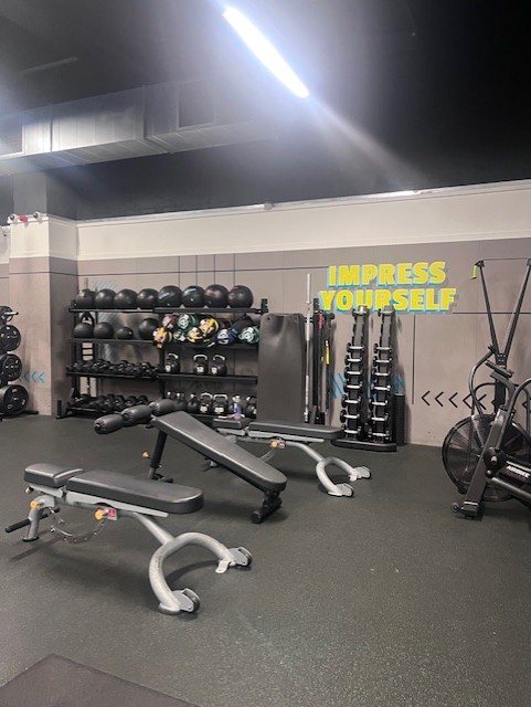 Modern gym interior with various exercise machines and equipment. Several people are present: one walking down the central aisle, two seated on benches using their phones, and others actively working out. The space features bright overhead lighting, gray flooring, and black ceiling panels.