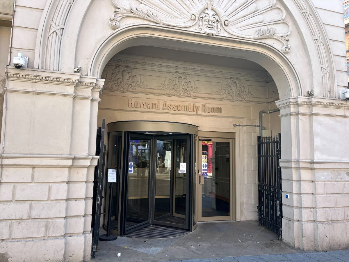 the entrance to the Howard Assembly Room, located within a grand, ornate stone building. The entrance features a glass revolving door beneath a decorative stone archway. Above the door 'Howard Assembly Room' is engraved into the stone wall.