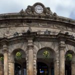 Leeds Corn Exchange-A grand, historic stone building with a curved facade and classical architectural elements. The structure features ornate carvings, arched windows, and decorative columns. A prominent round clock is set into a richly adorned pediment at the top center. Hanging flower baskets add vibrant greenery beneath the arches, softening the formal stonework.
