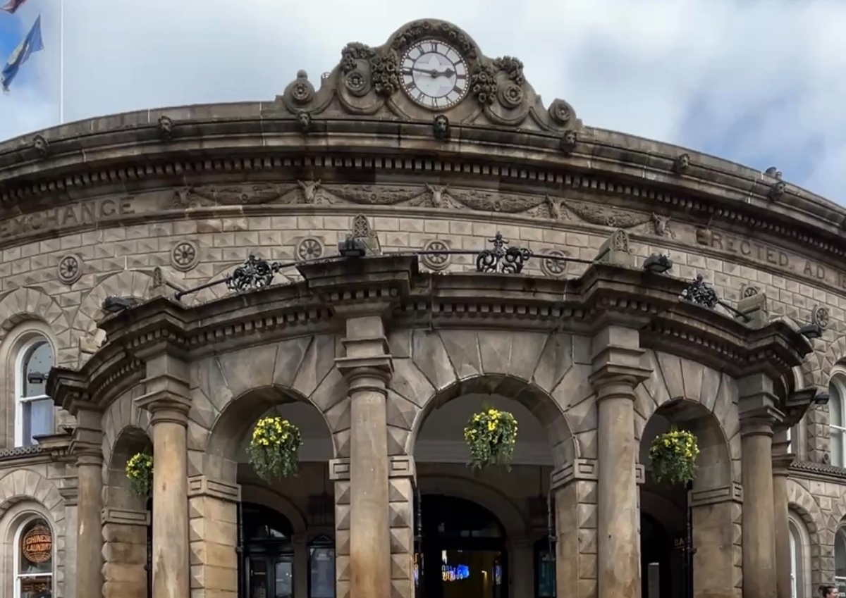 Leeds Corn Exchange-A grand, historic stone building with a curved facade and classical architectural elements. The structure features ornate carvings, arched windows, and decorative columns. A prominent round clock is set into a richly adorned pediment at the top center. Hanging flower baskets add vibrant greenery beneath the arches, softening the formal stonework.