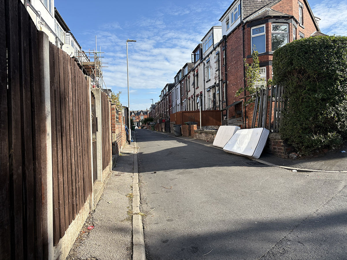 A quiet residential street with brick houses, wooden fences, and two discarded mattresses leaning against a stone wall on the right side of the road under a clear blue sky.