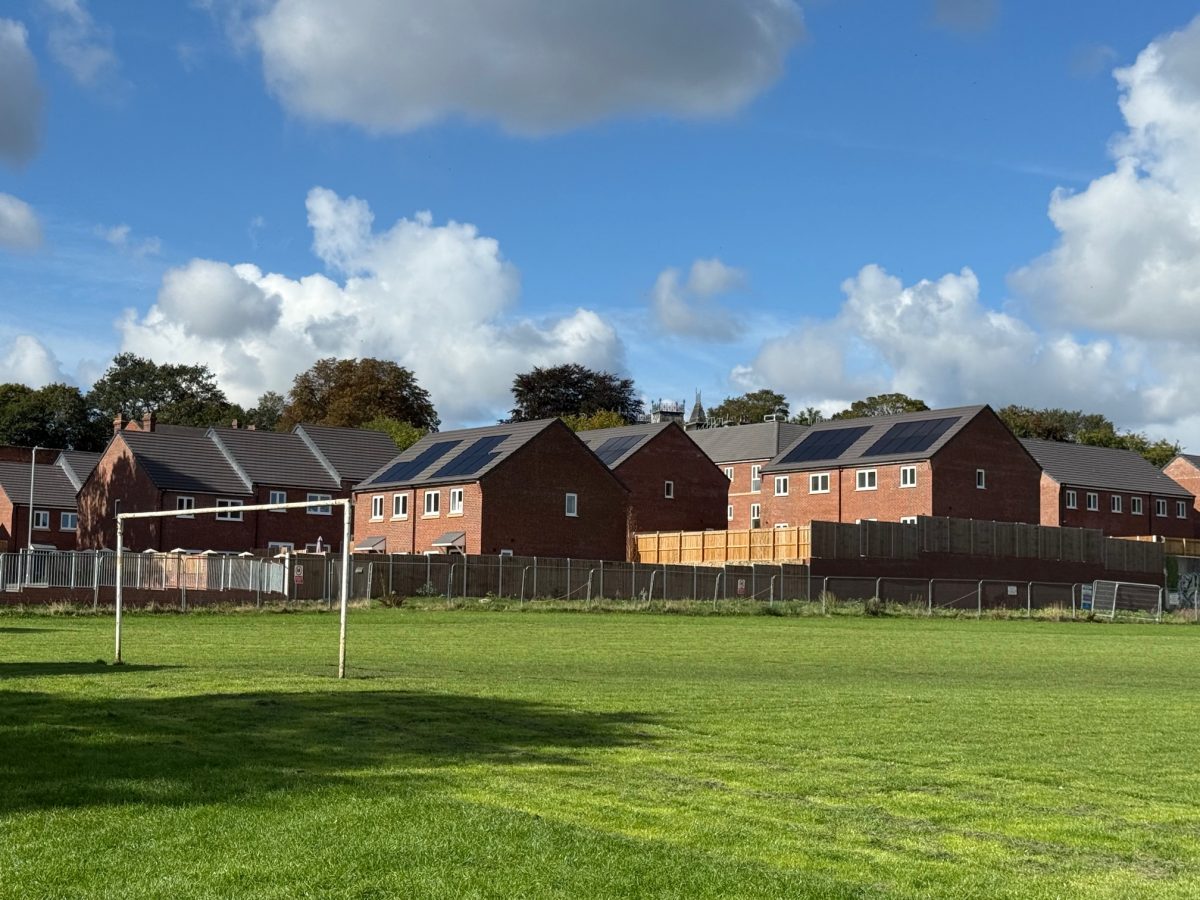 A row of newly built red-brick houses with dark grey tiled roofs, some equipped with solar panels, stands behind a wooden fence and temporary construction barriers. In the foreground, a grassy playing field with a worn football goalpost is visible. The sky is partly cloudy with patches of bright blue, casting natural light across the scene. Tall trees with early autumn foliage are visible behind the houses.