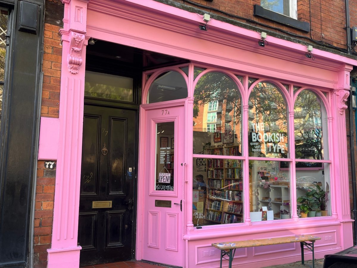 A bookstore with a bright pink facade and large windows displays "The Bookish Type" sign. The door reads "Open," and books are visible inside. A bench is outside the shop.
