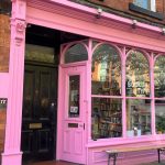 A bookstore with a bright pink facade and large windows displays "The Bookish Type" sign. The door reads "Open," and books are visible inside. A bench is outside the shop.