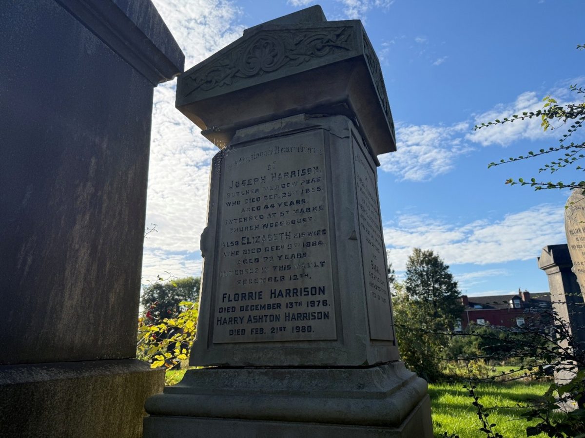 A photograph taken in a graveyard during daylight shows a tall, weathered stone grave monument inscribed with multiple names. The central inscription reads: "Joseph Harrison, butcher, Meadow Lane, who died Sept. 30th, 1855, aged 44 years. Interred at St. Mark’s Church Woodhouse." "Also Elizabeth, his wife, who died Dec. 1st, 1888, aged 77 years. Interred in this vault Dec. 4th." "Florrie Harrison, died December 13th, 1976." "Harry Ashton Harrison, died Feb. 21st, 1980." The monument features an ornate carved floral design at the top. Sunlight illuminates parts of the stone, creating shadows that emphasize its age and texture. In the background, trees and other gravestones are visible, along with a row of red-brick houses. The sky is bright with scattered clouds, suggesting a calm autumn or spring day. The foreground is partially framed by the edge of another grave monument and overhanging branches.