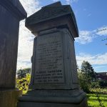 A photograph taken in a graveyard during daylight shows a tall, weathered stone grave monument inscribed with multiple names. The central inscription reads: "Joseph Harrison, butcher, Meadow Lane, who died Sept. 30th, 1855, aged 44 years. Interred at St. Mark’s Church Woodhouse." "Also Elizabeth, his wife, who died Dec. 1st, 1888, aged 77 years. Interred in this vault Dec. 4th." "Florrie Harrison, died December 13th, 1976." "Harry Ashton Harrison, died Feb. 21st, 1980." The monument features an ornate carved floral design at the top. Sunlight illuminates parts of the stone, creating shadows that emphasize its age and texture. In the background, trees and other gravestones are visible, along with a row of red-brick houses. The sky is bright with scattered clouds, suggesting a calm autumn or spring day. The foreground is partially framed by the edge of another grave monument and overhanging branches.