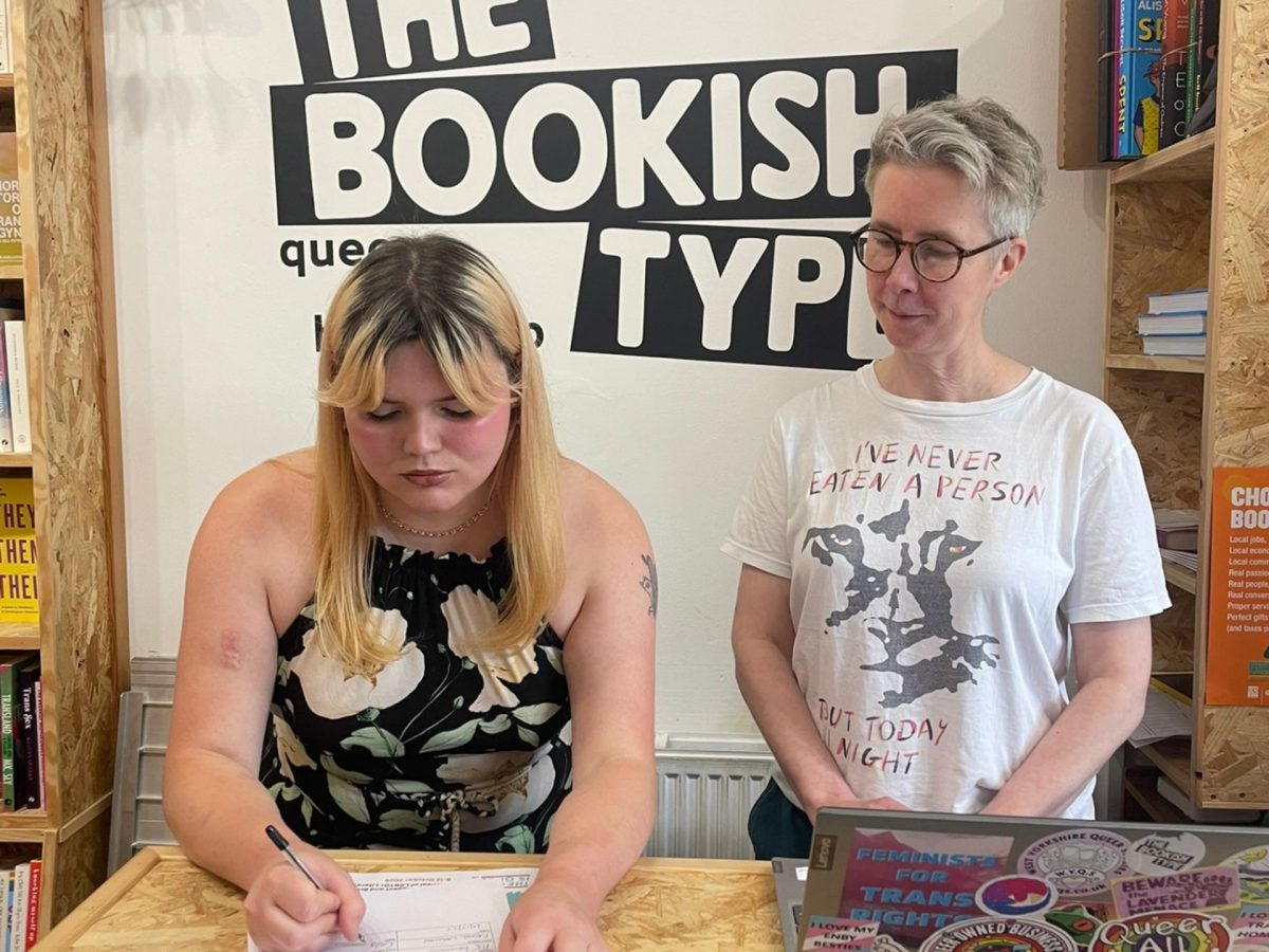 Two people stand at a counter in a bookstore; one is writing on a form while the other watches. Shelves with books and a sign reading "The Bookish Type" are visible in the background.