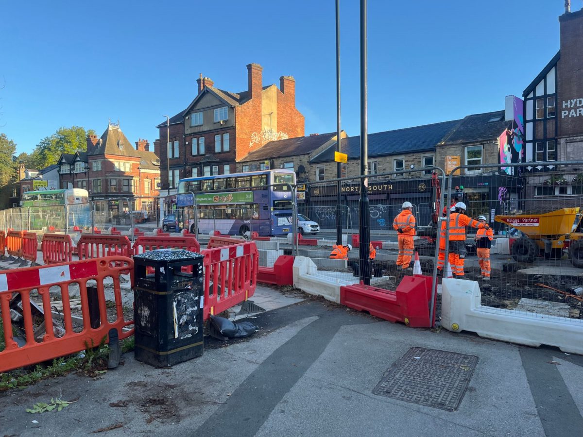 Road construction site in an urban area with several workers wearing orange high-visibility safety gear. Red and white barriers surround the site, and a digger and construction equipment are visible. In the background, buses are passing by on the road near buildings. The sky is clear and blue.