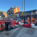 Road construction site in an urban area with several workers wearing orange high-visibility safety gear. Red and white barriers surround the site, and a digger and construction equipment are visible. In the background, buses are passing by on the road near buildings. The sky is clear and blue.