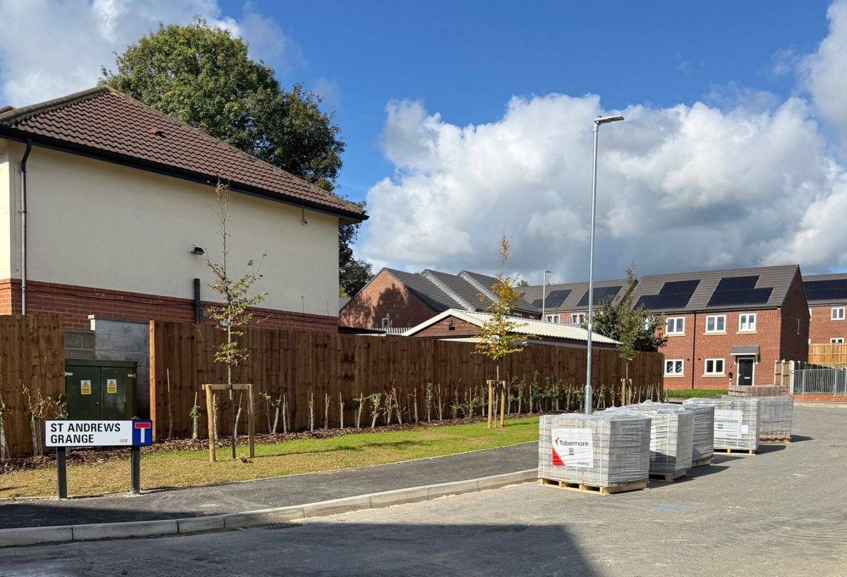 A newly built suburban street named "St Andrews Grange," marked by a road sign on the left. The street features modern brick houses with tiled roofs, some fitted with solar panels. Young trees are planted along a fenced boundary, supported by wooden stakes. On the right side of the road, several pallets of paving slabs branded "Tobermore" are placed, suggesting ongoing construction or landscaping work. The scene is under a bright blue sky with scattered clouds.