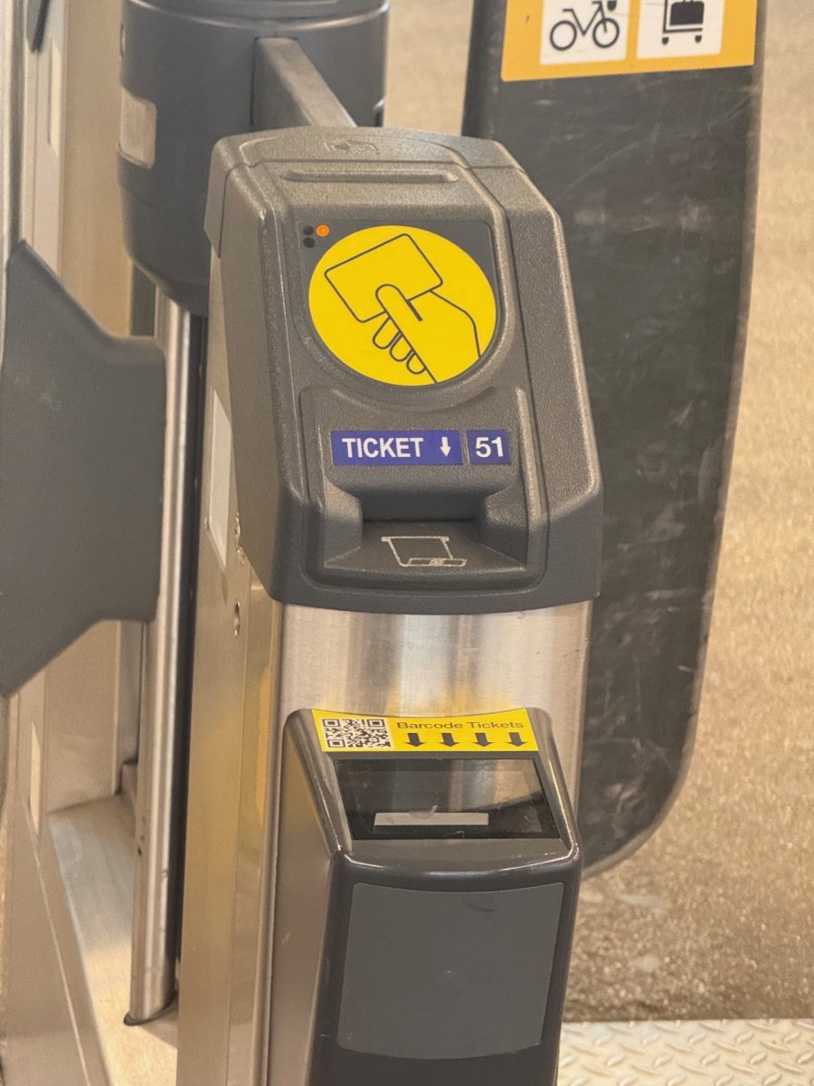 A close-up view of a ticket barrier at a train station. The machine has a yellow circular symbol showing a hand holding a card, indicating where passengers should tap their ticket or card. Below the symbol, a blue label reads “TICKET ↓ 51.” A barcode scanner slot is positioned lower down with a yellow sticker showing arrows and the words “Barcode Tickets.” The barrier arms are partially visible on either side of the machine.