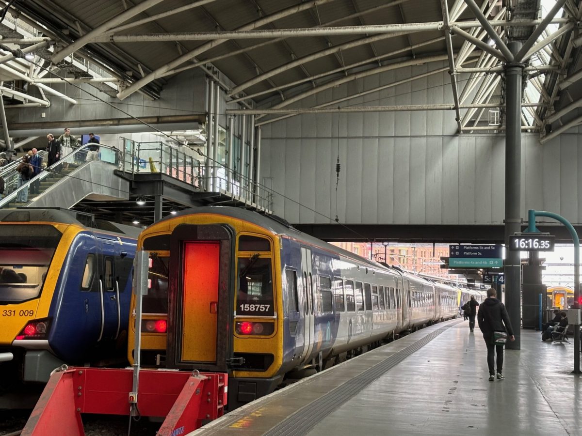 A busy indoor train station featuring two stationary trains at adjacent platforms. The train on the left is a modern Northern Class 331 electric unit, numbered 331009, with a sleek blue and yellow design. Next to it on the right is an older Northern Class 158 diesel unit, numbered 158757, with a more classic shape and similar blue and yellow livery. The station has a high, arched roof with exposed beams and industrial lighting. Passengers are visible on the platform and escalator, and a digital clock displays the time as 16:16:35. Overhead signs direct travellers to various platforms, and daylight is visible through the far end of the station.