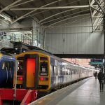 A busy indoor train station featuring two stationary trains at adjacent platforms. The train on the left is a modern Northern Class 331 electric unit, numbered 331009, with a sleek blue and yellow design. Next to it on the right is an older Northern Class 158 diesel unit, numbered 158757, with a more classic shape and similar blue and yellow livery. The station has a high, arched roof with exposed beams and industrial lighting. Passengers are visible on the platform and escalator, and a digital clock displays the time as 16:16:35. Overhead signs direct travellers to various platforms, and daylight is visible through the far end of the station.