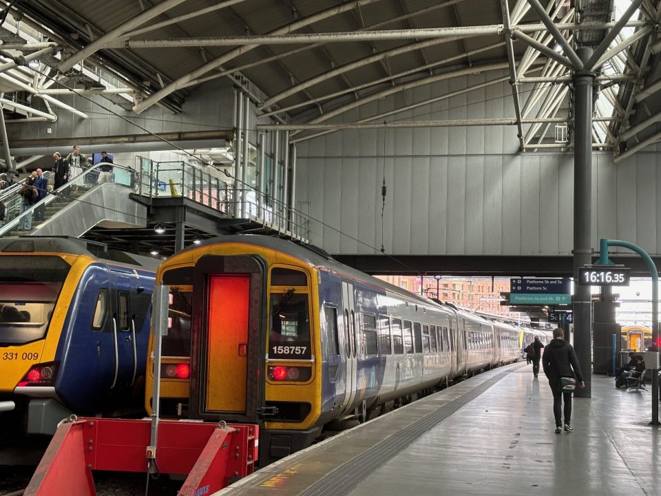 A busy indoor train station featuring two stationary trains at adjacent platforms. The train on the left is a modern Northern Class 331 electric unit, numbered 331009, with a sleek blue and yellow design. Next to it on the right is an older Northern Class 158 diesel unit, numbered 158757, with a more classic shape and similar blue and yellow livery. The station has a high, arched roof with exposed beams and industrial lighting. Passengers are visible on the platform and escalator, and a digital clock displays the time as 16:16:35. Overhead signs direct travellers to various platforms, and daylight is visible through the far end of the station.