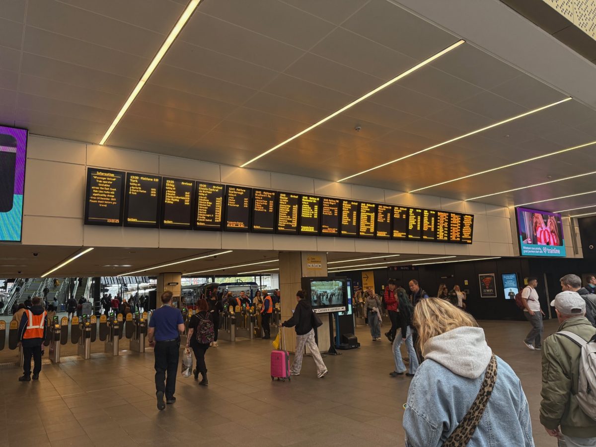 Interior of a busy train station concourse with a large digital departure board displaying upcoming train times, platforms, and destinations in bright yellow text on a black background. Below the board, passengers move through automated ticket barriers, while others walk around or check the screens. Several staff in high-visibility orange vests assist passengers. To the left, escalators lead up to the platforms, and on the right side, a large digital advertisement screen displays a Trainline promotion. The station is brightly lit with a modern ceiling featuring linear lights, and the atmosphere is bustling with activity.